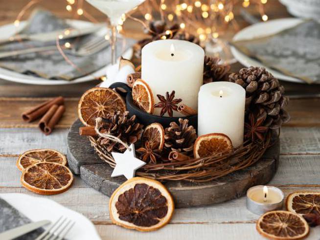 Varias velas blancas en un centro de mesa decoradas con piñas y frutas desecadas.