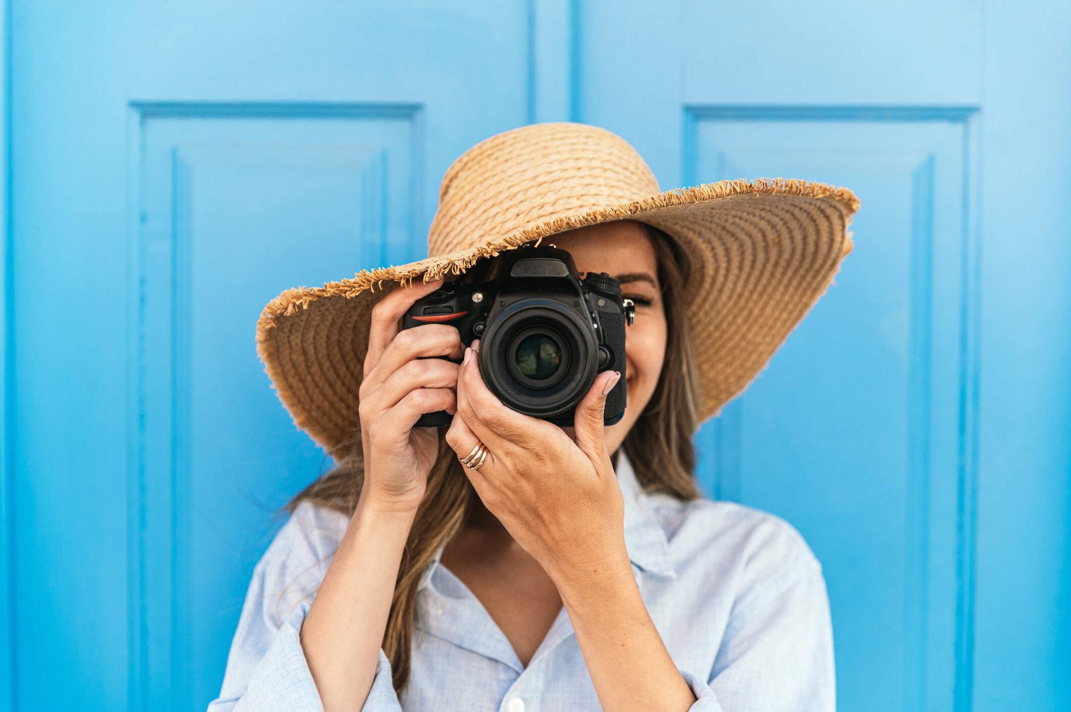 Female traveler in straw hat taking picture on professional photo camera while standing near blue building on street during summer vacation in Lagos in Portugal