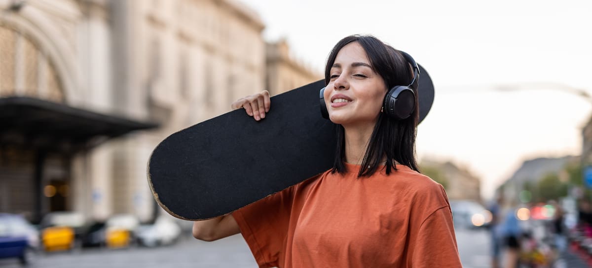 One young woman, a young Spanish woman with a skateboard on her shoulder walks down the street, dressed casually in an orange T-shirt