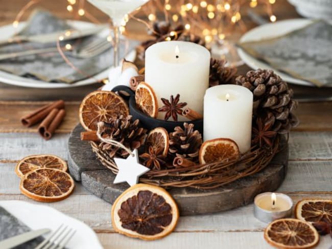 Varias velas blancas en un centro de mesa decoradas con piñas y frutas desecadas.