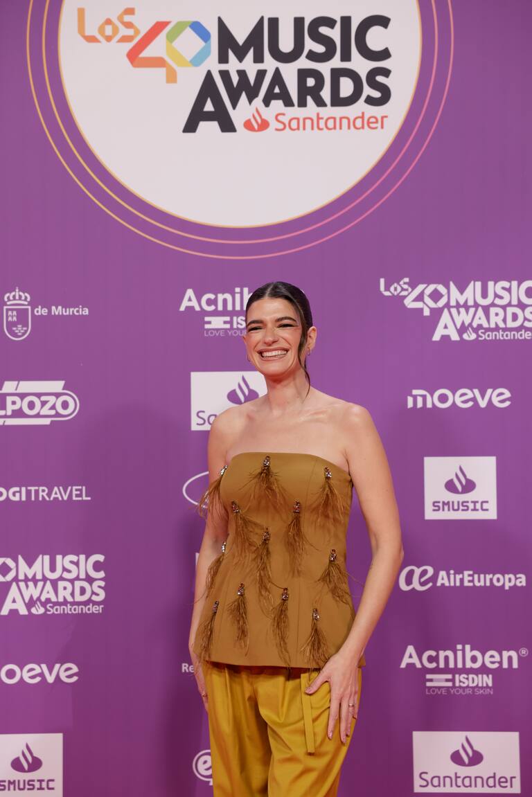 Bonbon Reich, la influencer, pasa por la alfombra de LOS40 Music Awards 2025 con un conjunto dos piezas en mostaza formado por un top escote palabra de honor con plumas que caen en forma de lágrima y un pantalón más sobrio del mismo color.